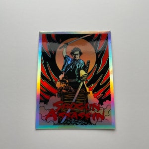 May include: A rectangular sticker with a holographic border featuring a graphic from the film "Shogun Assassin." The image shows a samurai with a sword raised, and another figure in a wooden box. The title is in red.