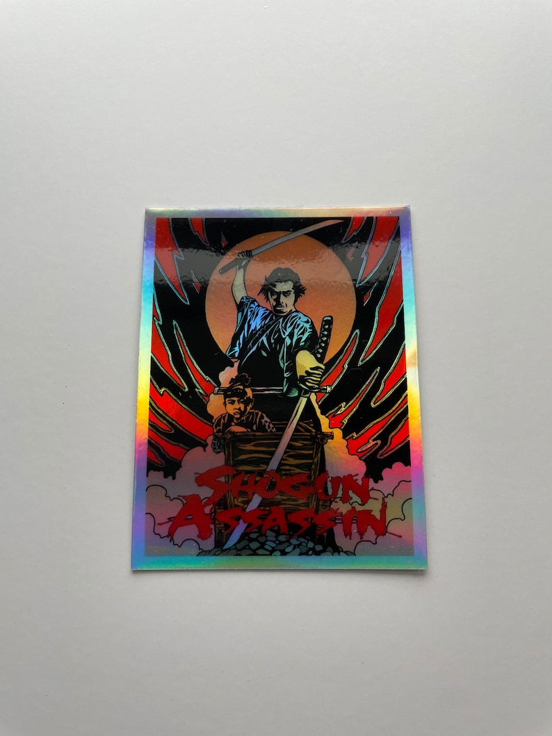May include: A rectangular sticker with a holographic border featuring a graphic from the film "Shogun Assassin." The image shows a samurai with a sword raised, and another figure in a wooden box. The title is in red.