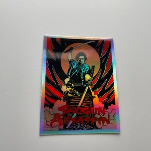 May include: A holographic sticker featuring a scene from Shogun Assassin. The artwork depicts a samurai wielding a sword, with a child in a cart. The background is a fiery red and black design with the movie title.