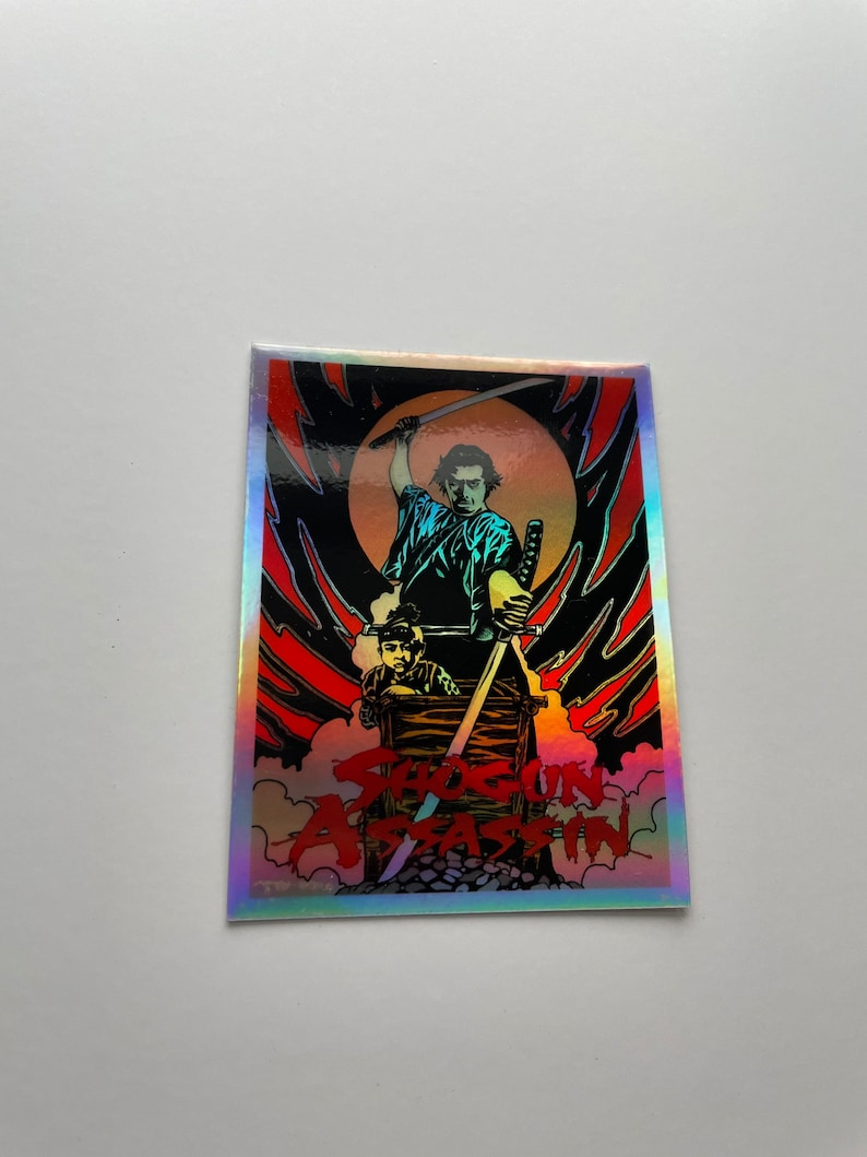 May include: A holographic sticker featuring a scene from Shogun Assassin. The artwork depicts a samurai wielding a sword, with a child in a cart. The background is a fiery red and black design with the movie title.