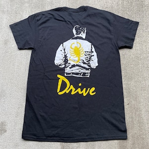 May include: Black t-shirt with a white and yellow graphic of a man wearing a jacket with a scorpion on the back. The text "Drive" is written in yellow below the graphic.