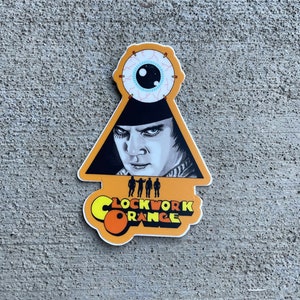 Puede incluir: Una pegatina amarilla y naranja con el texto "Clockwork Orange" y una imagen en blanco y negro de un hombre con un sombrero negro y un ojo grande encima de él.