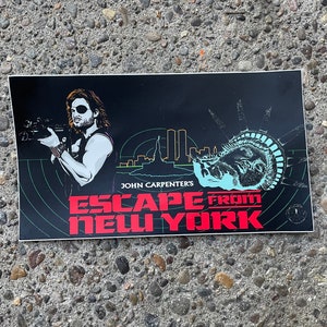May include: A black and white sticker with a graphic of a man holding a gun, a cityscape, and the Statue of Liberty. The text "JOHN CARPENTER'S ESCAPE FROM NEW YORK" is in red and white.