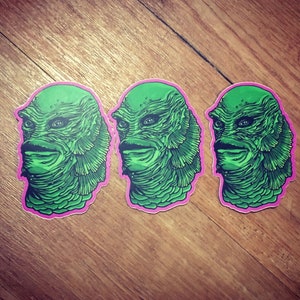 May include: Three green and pink stickers featuring a creature from the black lagoon. The creature has green skin, a wide mouth, and a toothy grin. The stickers are ideal for adding a touch of horror to any surface.