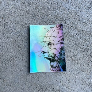 May include: A sticker featuring a black and white illustration of a person's face with a large number of pins protruding from their head. The background is a holographic iridescent blue and purple.