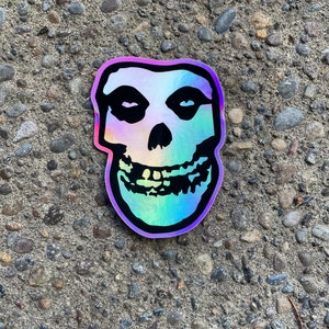 May include: A holographic sticker featuring the iconic Misfits skull logo. The skull has black eyes and a black nose, with a rainbow gradient effect across the face. The sticker is outlined in black and sits on a textured gray surface.