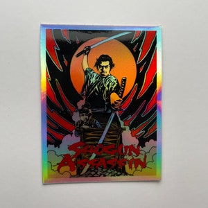 May include: Holographic sticker featuring a stylized illustration from "Shogun Assassin." The artwork depicts a samurai with two swords, set against a red and black background with a rising sun. The title is in red.