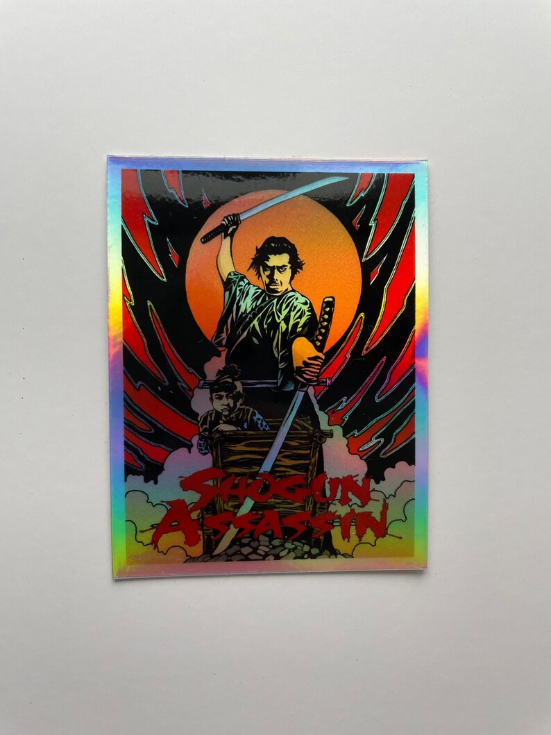 May include: Holographic sticker featuring a stylized illustration from "Shogun Assassin." The artwork depicts a samurai with two swords, set against a red and black background with a rising sun. The title is in red.