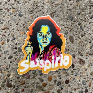 May include: A colorful sticker featuring a woman with long dark hair and a bright blue and yellow face. She is holding a knife and has a menacing expression. The sticker has the word "Suspiria" written in white letters on a yellow background.