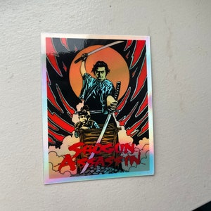 May include: A holographic sticker featuring a stylized illustration from "Shogun Assassin." The artwork depicts a samurai figure with a raised sword, set against a red and black background with the film's title.