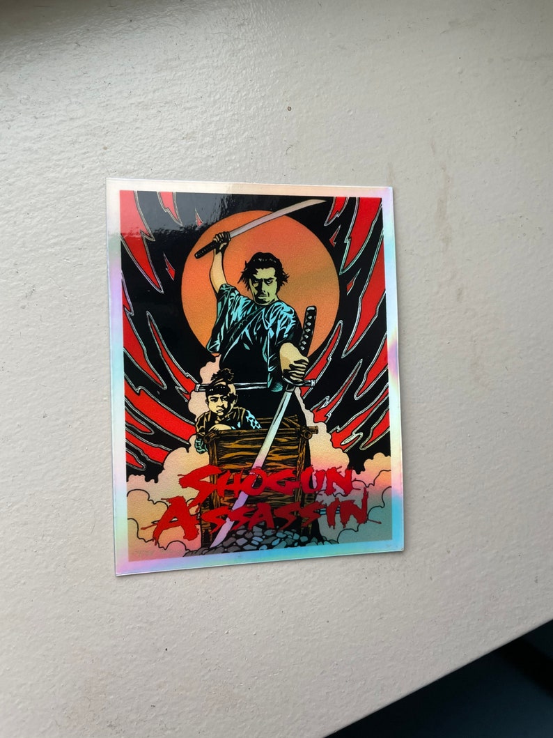 May include: A holographic sticker featuring a stylized illustration from "Shogun Assassin." The artwork depicts a samurai figure with a raised sword, set against a red and black background with the film's title.