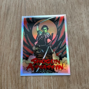 May include: Holographic sticker featuring a stylized illustration of a samurai wielding two swords. The image includes a figure in a wooden cart and the text "Shogun Assassin" in red, with a black and red background.