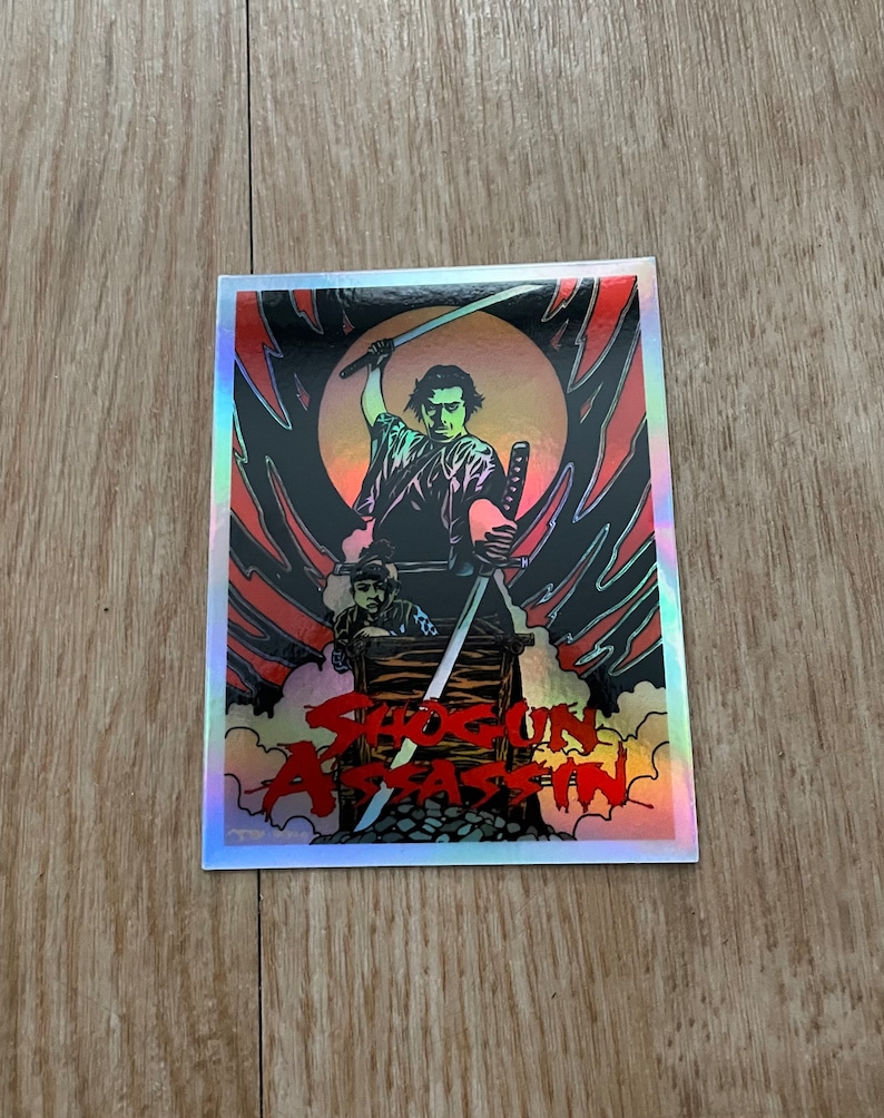 May include: Holographic sticker featuring a stylized illustration of a samurai wielding two swords. The image includes a figure in a wooden cart and the text "Shogun Assassin" in red, with a black and red background.