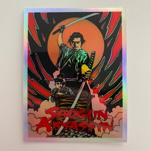 May include: Holographic sticker featuring a stylized illustration from the film "Shogun Assassin." The artwork depicts a samurai with a raised sword, a child, and the film's title in red. The background includes a red and black design with a yellow sun.