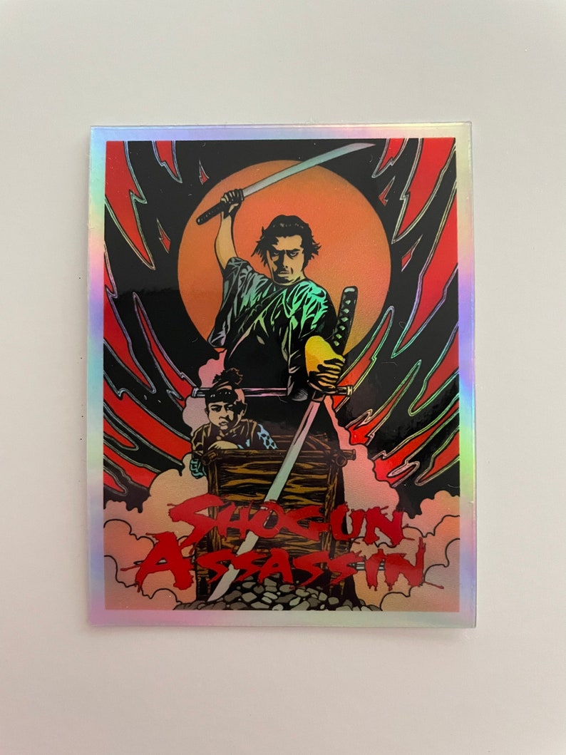 May include: Holographic sticker featuring a stylized illustration from the film "Shogun Assassin." The artwork depicts a samurai with a raised sword, a child, and the film's title in red. The background includes a red and black design with a yellow sun.