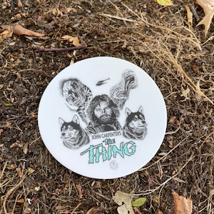 May include: A white, round coaster featuring a black and white graphic from John Carpenter's "The Thing." The design includes images of characters and the film's title in a teal font. The coaster is resting on a bed of soil and leaves.