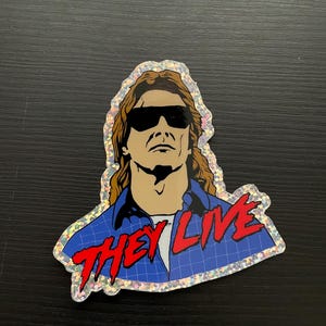 May include: A glittery sticker featuring a man with long brown hair wearing sunglasses and a blue plaid shirt. The text "THEY LIVE" is written in red letters below the man.