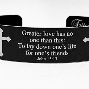 Könnte beinhalten: Schwarzes Metallarmband mit einem weißen Kreuz auf jeder Seite und der Inschrift "Greater love has no one than this: To lay down one's life for one's friends John 15:13".