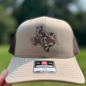 May include: A tan and brown trucker hat with a camouflage Texas state outline patch and a white star. The hat has a black mesh back and a Richardson 112 logo.