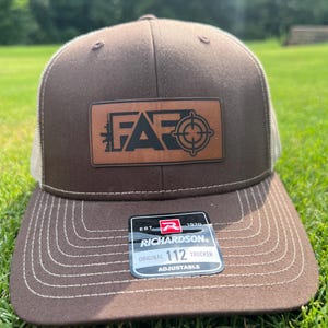 Puede incluir: Gorra de camionero marrón y blanca con un parche de cuero marrón que dice "FAR" con un símbolo de diana. La gorra es ajustable y tiene una etiqueta Richardson 112.