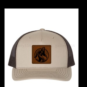 May include: A brown and black trucker hat with a brown leather patch featuring a black silhouette of Bigfoot. The patch is stitched onto the front of the hat.