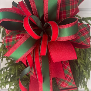 May include: A decorative bow featuring a mix of solid red velvet, solid dark green satin, and red and green plaid fabric ribbons. The bow has multiple loops and long trailing tails, with some edges accented by red glitter, attached to greenery.