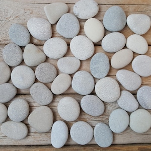 Può includere: Una collezione di ciottoli di fiume lisci, grigi e biancastri di varie dimensioni e forme. Le pietre sono disposte su una superficie di legno marrone chiaro invecchiato, creando un'estetica naturale e terrosa. Le pietre variano dal grigio chiaro al biancastro.