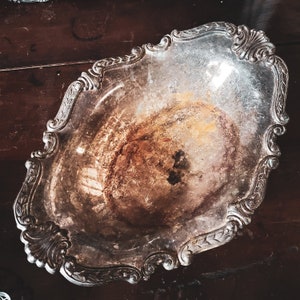 May include: A silver serving dish with an ornate design and a tarnished finish. The dish is oval shaped and has a raised rim.