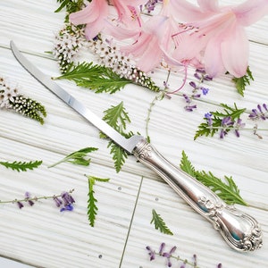 May include: A silver butter knife with an ornate handle, lying on a white wooden surface with pink and purple flowers scattered around it.