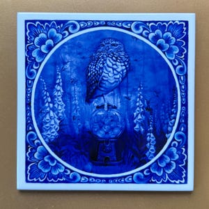 Może przedstawiać: Niebiesko-biała płytka ceramiczna z ozdobnym, kwiatowym obramowaniem. W centrum znajduje się niebiesko-biała ilustracja sowy siedzącej na maszynie do gum do żucia, otoczonej naparstniczkami i kolibrami.
