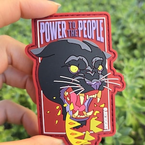 May include: A rectangular patch featuring a black panther design with yellow eyes and a purple mouth. The patch has the words "POWER TO THE PEOPLE" in purple text. The border is red.