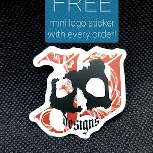 Puede incluir: Una pegatina de calavera roja y negra con el texto "JD Designs" sobre un fondo negro. La pegatina anuncia una pegatina de logotipo mini gratuita con cada pedido.