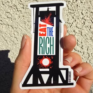May include: Black and white sticker of a guillotine with red blood splatter. The sticker says "Eat the Rich" in green and white text. The sticker is on a white background and has the text "@JDESIGNISART" in the bottom right corner.