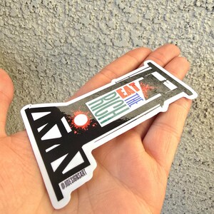 May include: A black and white sticker with a guillotine design. The sticker says "Eat the Rich" in colorful letters. The sticker is held in a hand.