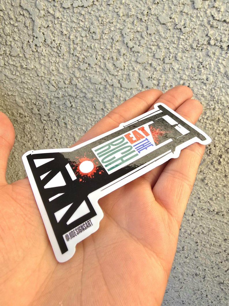 May include: A black and white sticker with a guillotine design. The sticker says "Eat the Rich" in colorful letters. The sticker is held in a hand.