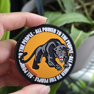 Puede incluir: Un parche redondo negro con el texto "ALL POWER TO THE PEOPLE" que rodea una pantera negra sobre un fondo naranja. La pantera gru&ntilde;e con dientes rojos. El parche tiene el texto "@IDESIGNSART".