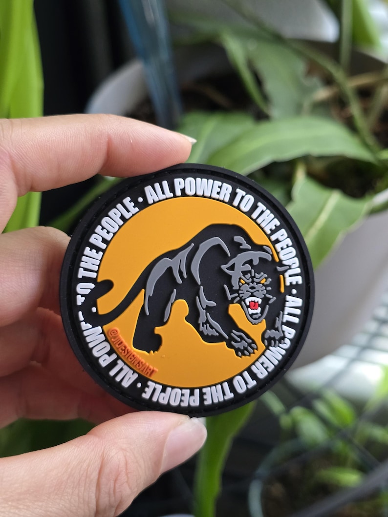 Puede incluir: Un parche redondo negro con el texto "ALL POWER TO THE PEOPLE" que rodea una pantera negra sobre un fondo naranja. La pantera gru&ntilde;e con dientes rojos. El parche tiene el texto "@IDESIGNSART".