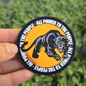 Puede incluir: Un parche redondo con una ilustraci&oacute;n de una pantera negra sobre un fondo amarillo. El texto blanco "ALL POWER TO THE PEOPLE" rodea el dise&ntilde;o. La pantera tiene detalles grises y dientes rojos, con un borde negro.