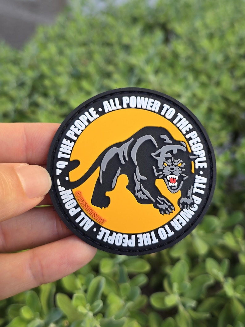 Puede incluir: Un parche redondo con una ilustraci&oacute;n de una pantera negra sobre un fondo amarillo. El texto blanco "ALL POWER TO THE PEOPLE" rodea el dise&ntilde;o. La pantera tiene detalles grises y dientes rojos, con un borde negro.