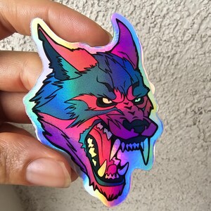 May include: A holographic sticker of a wolf's head with an angry expression. The wolf's fur is a gradient of blue, purple, and red.