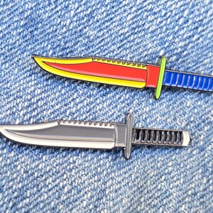 May include: Two enamel pins shaped like survival knives. The top pin features a bright red blade with a yellow edge, a lime green guard, and a blue handle. The bottom pin is finished in a metallic silver color with a dark grey blade and handle.