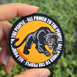 Puede incluir: Parche redondo negro con una ilustraci&oacute;n de pantera negra sobre fondo naranja. El texto blanco "ALL POWER TO THE PEOPLE" rodea el borde. El parche se sostiene en una mano, sobre un fondo verde borroso.
