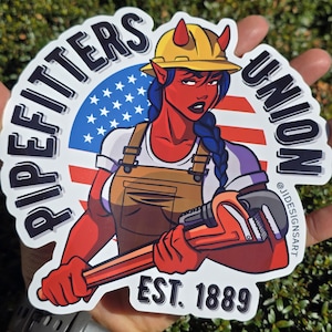 Puede incluir: Una pegatina con un personaje de piel roja con cuernos, que lleva un casco amarillo y sostiene una llave inglesa. El personaje está situado frente a una bandera americana. También están presentes las palabras "PIPEFITTERS UNION" y "EST. 1889".