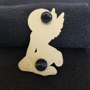 Puede incluir: Pin de metal dorado de un &aacute;ngel arrodillado con alas. El pin tiene dos tapones de goma negra en la parte posterior.