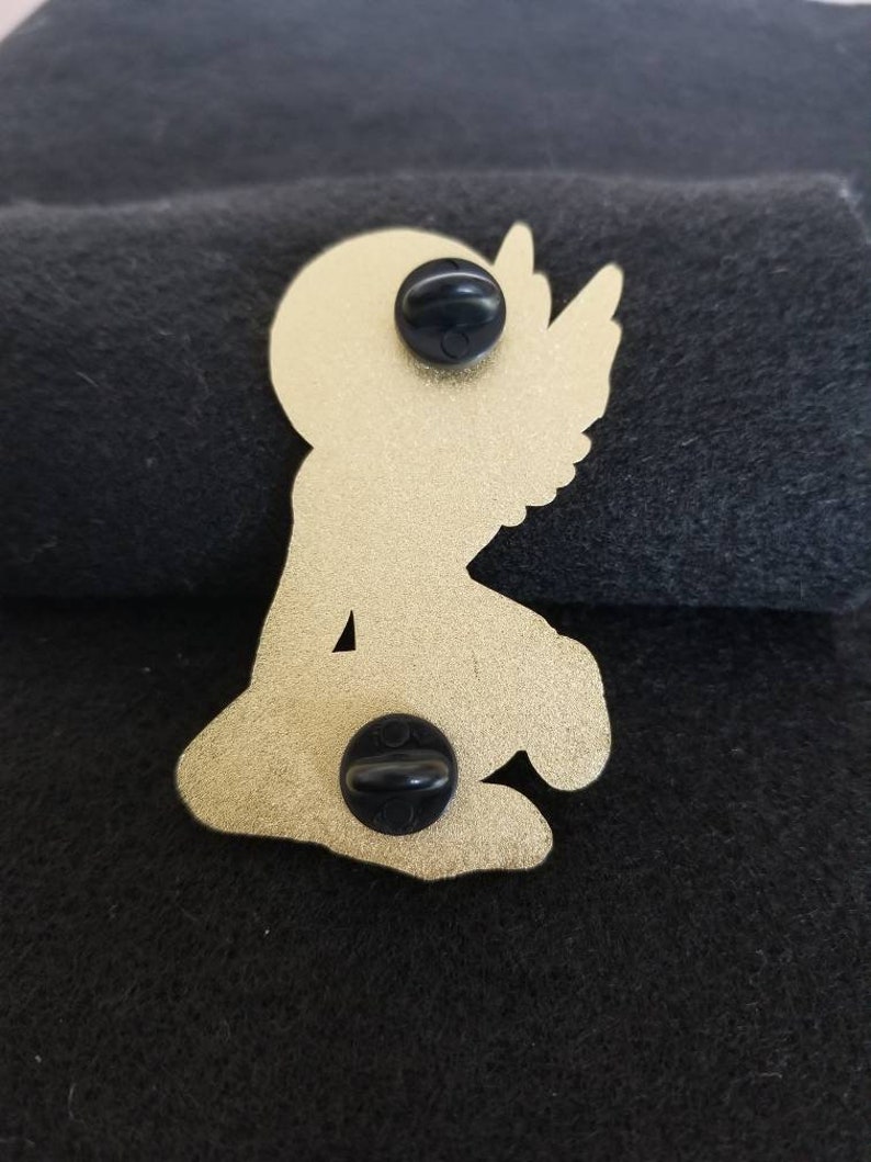 Puede incluir: Pin de metal dorado de un &aacute;ngel arrodillado con alas. El pin tiene dos tapones de goma negra en la parte posterior.