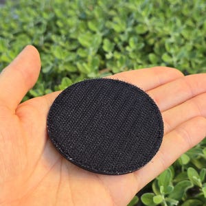 Puede incluir: Un parche redondo negro con una superficie texturizada, sostenido en una mano abierta. El parche est&aacute; hecho de un material tejido y tiene un respaldo de velcro. El fondo es follaje verde borroso.