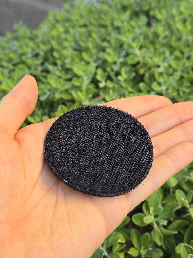 Puede incluir: Un parche redondo negro con una superficie texturizada, sostenido en una mano abierta. El parche est&aacute; hecho de un material tejido y tiene un respaldo de velcro. El fondo es follaje verde borroso.