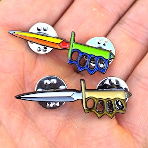 May include: Two metal enamel pins shaped like daggers with integrated brass knuckles. The top pin features a yellow, orange, red, green, and blue color scheme. The bottom pin is colored in shades of silver, black, and muted gold with metal backings.