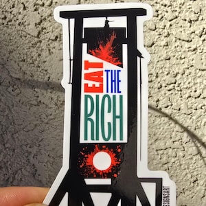 May include: Black and white sticker of a guillotine with red blood splatter and the text "Eat the Rich" in red, green, and blue. The sticker has a white border and the text "@JIDESIGNART" in the bottom right corner.