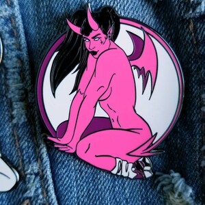 Puede incluir: Pin de esmalte rosa de una mujer demonio con alas, sentada con las piernas cruzadas. El pin est&aacute; sobre un fondo de mezclilla.